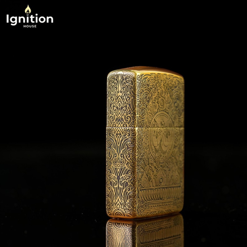 May include: A gold-colored lighter with intricate engraved patterns. The lighter has a rectangular shape with rounded edges and a hinged lid. The design includes floral and geometric motifs. The brand name "Ignition House" is in the top left corner.