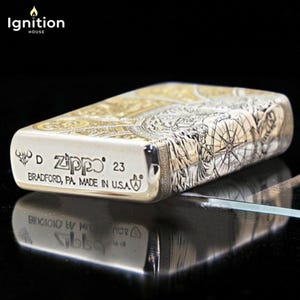 May include: A silver Zippo lighter with intricate engraved designs. The lighter features a compass rose and other detailed artwork. The text "Zippo 23 Bradford, PA. Made in U.S.A." is visible on the front. The lighter is displayed on a reflective surface.