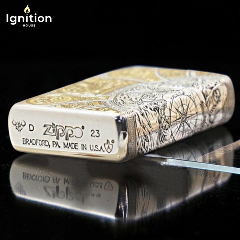 May include: A silver Zippo lighter with intricate engraved designs. The lighter features a compass rose and other detailed artwork. The text "Zippo 23 Bradford, PA. Made in U.S.A." is visible on the front. The lighter is displayed on a reflective surface.