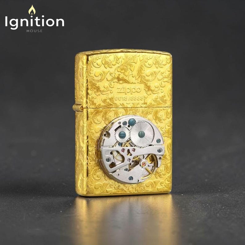 Puede incluir: Un encendedor Zippo dorado con un intrincado dise&ntilde;o floral y un mecanismo de reloj visible. El encendedor presenta el logotipo de Zippo y el n&uacute;mero de serie. El logotipo de Ignition House est&aacute; en la esquina superior izquierda.