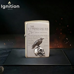 May include: A silver-toned metal lighter featuring an embossed raven perched on a skull. The front surface displays the text THE EXPENDABLES, CHOOSE YOUR WEAPON, and a poem that reads: This is how we face the death, we can not change, only slowly dilute it, the dark and more swallow us! more close to us!