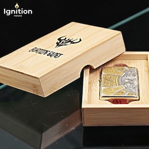 May include: A decorative lighter with a gold and silver design, presented in a wooden box. The lighter features an ornate pattern and a detailed image. The box is made of light-colored wood and has the text "BURNING GAINS".