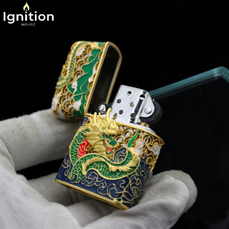 May include: An open, ornate lighter with a dragon design. The lighter features a gold dragon with green and red accents on a blue background. The lid and body have a gold and green decorative pattern. The brand "Ignition House" is in the top left corner.