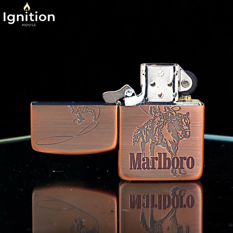 May include: A copper-colored metal lighter with the word "Marlboro" and a cowboy on a horse etched on the front. The lighter is open, revealing the interior mechanism. The "Ignition House" logo is in the top left corner.