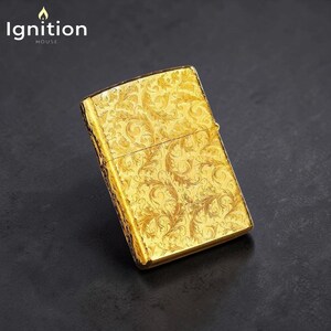 Puede incluir: Un encendedor dorado con un intrincado dise&ntilde;o floral. El encendedor, de forma rectangular, tiene una tapa con bisagras. El texto "Ignition House" est&aacute; en la esquina superior izquierda. El encendedor est&aacute; sobre una superficie oscura.