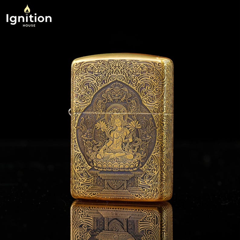 May include: A gold-colored metal lighter with an ornate engraved design. The design features a seated figure with lotus flowers, surrounded by intricate patterns. The text "Ignition House" is in the upper left corner.