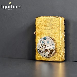 Puede incluir: Un encendedor Zippo dorado con un intrincado dise&ntilde;o floral y un mecanismo de reloj visible. El encendedor tiene la palabra "Zippo" y otro texto grabado. El logotipo de "Ignition House" est&aacute; en la esquina superior izquierda.