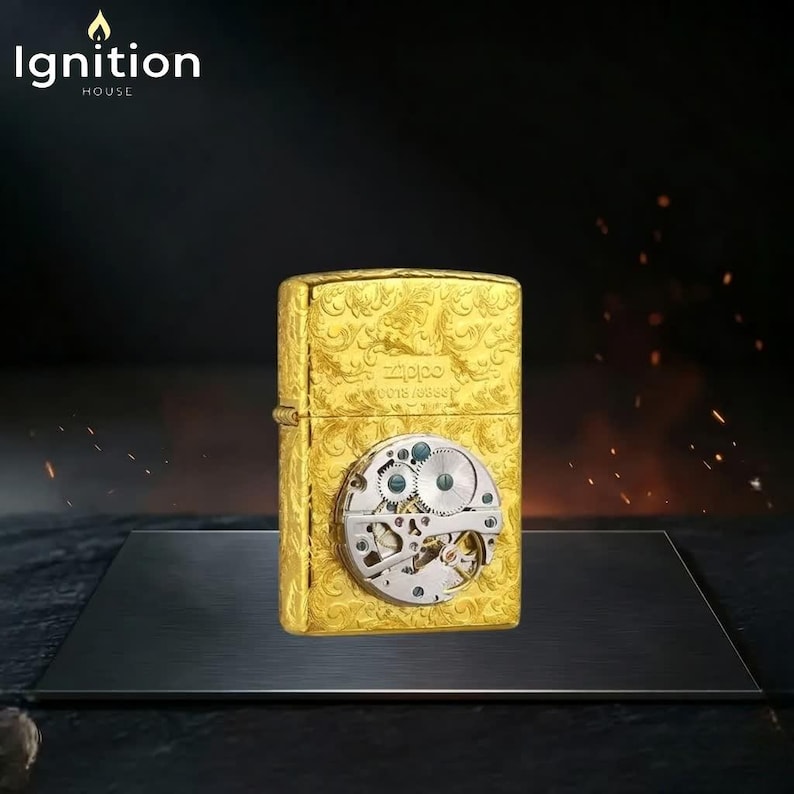 Puede incluir: Un encendedor Zippo dorado con un intrincado dise&ntilde;o floral y un mecanismo de reloj visible. El encendedor tiene el nombre de la marca y el n&uacute;mero de serie grabados. La imagen tambi&eacute;n incluye el texto "Ignition House".