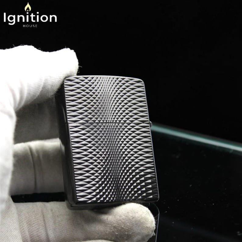 May include: Black and silver lighter with a geometric wave pattern. The lighter has a rectangular shape and a hinged lid. The text "Ignition House" is in the upper left corner. The lighter is held by a gloved hand.