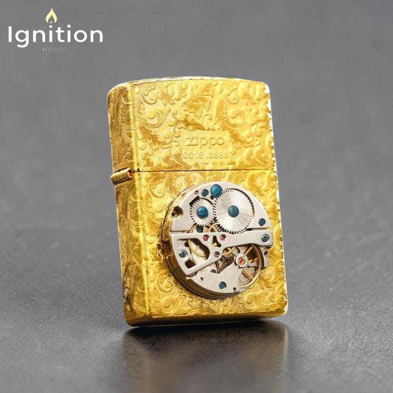 Puede incluir: Un encendedor Zippo dorado con un intrincado dise&ntilde;o floral y un mecanismo de reloj visible. El encendedor presenta el logotipo y el texto de Zippo, con una vista detallada de los engranajes internos y acentos azules. La palabra "Ignition" est&aacute; en la esquina superior izquierda.