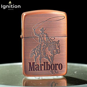 May include: A copper-colored metal lighter with a cowboy roping a horse design. The word "Marlboro" is printed in a dark brown color on the bottom of the lighter. The top left corner has the text "Ignition House".