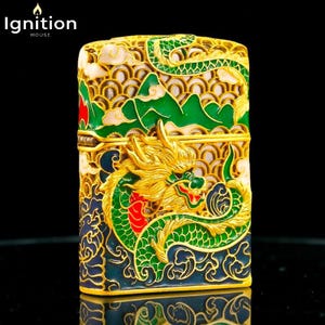 May include: Ornate gold-colored lighter with a detailed dragon design. The dragon is green and gold, set against a backdrop of green mountains and blue waves. The lighter has the text "Ignition House" in the upper left corner.