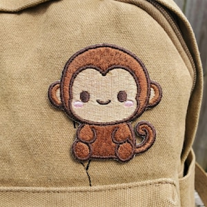 May include: A tan canvas backpack featuring a centered embroidered patch of a brown monkey. The monkey has a light beige face, dark brown ears, a small curved smile, and soft pink blush marks on its cheeks, with a curled tail to the right side.