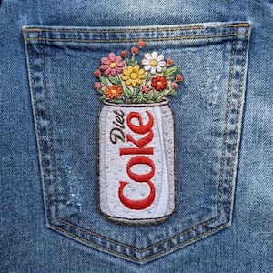 May include: A blue denim pocket featuring an embroidered patch of a silver soda can labeled Diet Coke in red text. The can is filled with a colorful bouquet of embroidered flowers in shades of pink, yellow, white, and orange with green leaves.