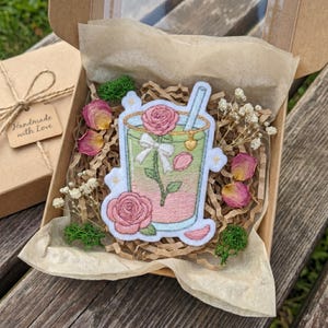 May include: An embroidered patch featuring a glass filled with a pink and green liquid, a pink rose, a white bow, and a blue straw with a small gold heart charm. The patch is nestled in a cardboard gift box with brown paper filler and dried flowers.