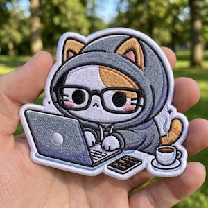 May include: An embroidered patch featuring a calico cat wearing a gray hoodie and black glasses, typing on a laptop. The cat is positioned next to a smartphone and a steaming cup of coffee. The design uses white, gray, orange, and black thread.