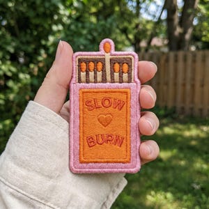 May include: An embroidered patch shaped like a matchbox with a pink border. The center is orange with the text SLOW BURN and a heart icon. The top section shows brown matchsticks with orange tips against a dark brown background.