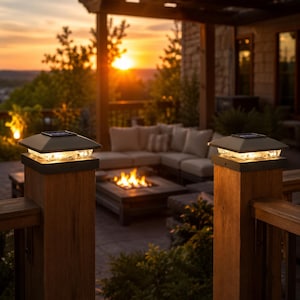 May include: Two square solar post cap lights with dark frames and clear glass panels are mounted on top of wooden deck posts. The lights emit a warm glow, illuminating the top of the posts while the sun sets in the background behind a patio.