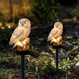 Puede incluir: Dos luces solares de jardín con forma de búho. Cada luz presenta un búho blanco posado sobre una base de madera artificial, con un brillo cálido. Las luces están montadas en estacas negras para el jardín.