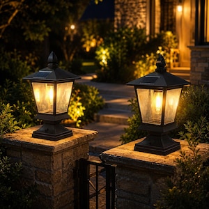 May include: Two black solar-powered post cap lights with a square base and a pointed finial top. Each lantern features clear glass panels and an internal warm-toned light bulb, mounted on top of stone pillars in an outdoor garden setting at night.