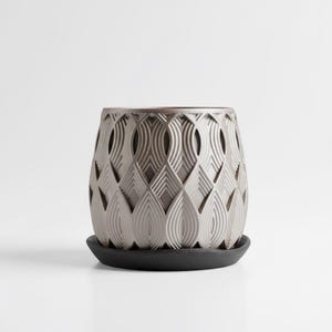 Könnte beinhalten: Grauer Keramik-Blumentopf mit einem geometrischen Muster aus sich überlappenden Blattformen. Der Topf steht auf einer runden, schwarzen Untertasse. Der Topf hat eine strukturierte Oberfläche und ein modernes Design.