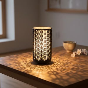 Könnte beinhalten: Zylindrische schwarze Metalllampe mit geometrischem Muster, die komplizierte Schatten auf einen Holztisch wirft. Die Lampe ist beleuchtet und erzeugt eine warme Atmosphäre. Eine kleine Schale und Blumen befinden sich ebenfalls auf dem Tisch.