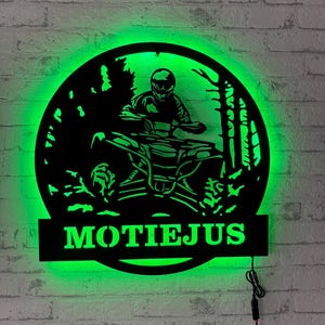 Könnte beinhalten: Ein rundes Wand-Schild aus schwarzem Metall mit der Silhouette einer Person auf einem Quad in einem Wald, beleuchtet durch hellgrünes LED-Licht. Der Name MOTIEJUS ist in großen Druckbuchstaben unten ausgeschnitten und leuchtet grün.