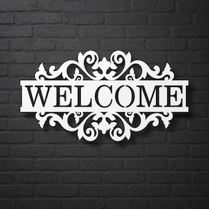 Könnte beinhalten: Weißes Metall-Willkommensschild mit kunstvollen Verzierungen. Das Wort „WELCOME“ ist in fetten Großbuchstaben ausgeschnitten. Das Schild ist an einer dunkelgrauen Backsteinmauer angebracht und erzeugt einen auffälligen Kontrast.
