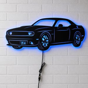 Pode incluir: Arte de parede preta em forma de carro com iluminação LED azul. O carro tem uma placa de matrícula que diz "A4E3". A arte é montada numa parede de tijolo branco e está ligada a uma fonte de alimentação através de um cabo preto.