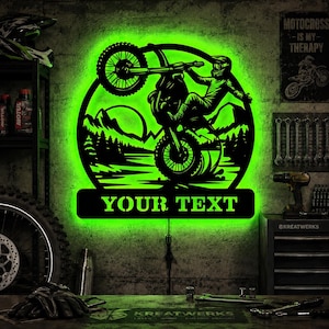 Könnte beinhalten: Ein rundes Wandbild aus schwarzem Metall mit einer grün hinterleuchteten LED-Silhouette eines Motocross-Fahrers, der einen Wheelie vor einer Berg- und Waldlandschaft macht, inklusive eines rechteckigen Sockels mit dem Text YOUR TEXT.