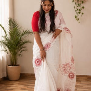 May include: A white saree featuring red block-printed motifs, including the Om symbol, a swastika, and a seated figure. The garment is paired with a solid red short-sleeved blouse, creating a contrast between the white fabric and red patterns.