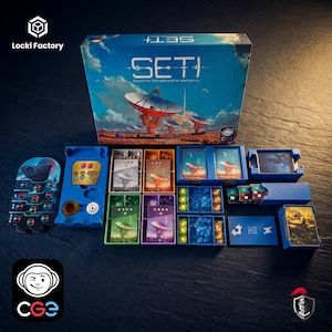 Peut inclure: Le jeu de société SETI: Search for Extraterrestrial Intelligence comprend une grande boîte bleue illustrée de paraboles, accompagnée de divers organisateurs en plastique bleu, vert, orange et violet contenant des cartes, jetons et marqueurs.
