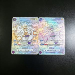 Könnte beinhalten: Zwei holografische Sammelkarten mit japanischem Text. Jede Karte zeigt eine Illustration eines Charakters in weißer Kleidung, umgeben von aufwendigen Mustern. Die Karten zeigen Werte von 10 und 12000.