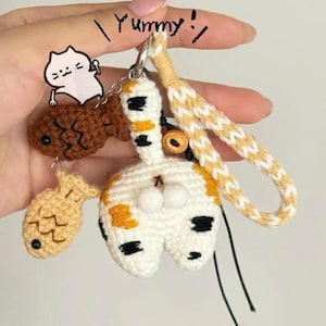 Op de afbeelding: Een handgemaakte gehaakte sleutelhanger met een witte kat met oranje en zwarte vlekken, en twee visbedels. De sleutelhanger heeft een gevlochten band in wit en geel. Het woord "Yummy!" staat erboven.