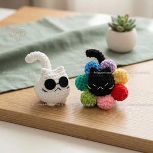 May include: Two crocheted cat figures. One is white with black sunglasses, the other is black with colorful petals. The figures are handmade and sit on a wooden surface. A small succulent in a white pot is in the background.