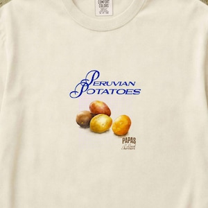 May include: An ivory short-sleeve t-shirt featuring a graphic of three potatoes. The text reads Peruvian Potatoes in blue script and Papas in smaller black font. The shirt has a crew neckline and a Comfort Colors brand label at the inner collar.