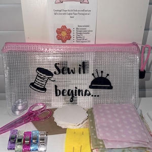 May include: A clear, rectangular zippered pouch with pink trim and the black text "Sew it begins..." featuring a spool and pincushion graphic. The kit includes pink scissors, white hexagon paper pieces, five colorful clips, two needles, and fabric squares.