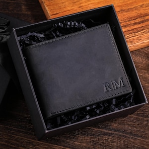 Puede incluir: Una cartera de cuero negro con las iniciales "RM" grabadas en la parte delantera, presentada en una caja de regalo negra con papel triturado. La caja está abierta, revelando la cartera. El fondo es una superficie de madera.
