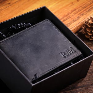 Könnte beinhalten: Schwarzes Lederportemonnaie mit den Initialen „RM“ auf der Vorderseite, präsentiert in einer schwarzen Geschenkbox. Das Portemonnaie liegt auf einem Holzgrund, mit einem Tannenzapfen im Hintergrund.