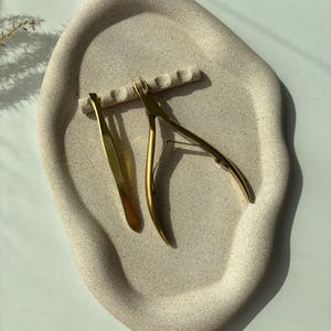 Könnte beinhalten: Eine beige, organisch geformte Schale mit strukturierter Oberfläche. Goldfarbene Nagelknipser und eine Pinzette liegen auf der Schale. Die Schale hat einen kleinen, erhöhten Bereich zur Aufnahme der Werkzeuge. Der Hintergrund ist eine weiche, neutrale Farbe.