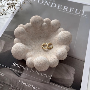 May include: A beige, speckled, circular trinket dish with a textured, bubble-like rim. The dish has a rough, stone-like finish and contains two small, plain gold-colored metal rings resting in the center of the bowl-shaped interior.