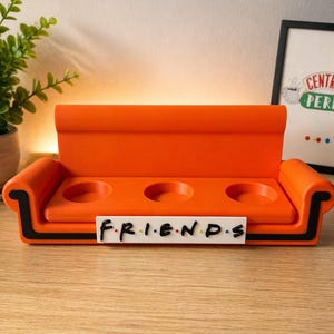 Può includere: Un oggetto arancione a forma di divano a tre fori con la parola "FRIENDS" in lettere nere su una base rettangolare bianca. Il divano ha finiture nere e si trova su una superficie di legno.