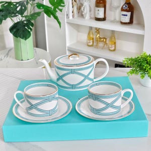 Puede incluir: Un juego de té de porcelana blanca que incluye una tetera y dos tazas con sus platos a juego. Cada pieza está decorada con un patrón repetitivo de cintas de color azul claro y un fino borde dorado. El conjunto se presenta sobre una caja azul.