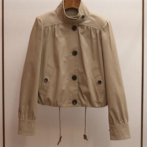 Puede incluir: Chaqueta beige con cuello alto y cierre de botones. La chaqueta tiene mangas largas con puños abotonados, dos bolsillos delanteros y un cordón en el dobladillo. Colgada de una percha de madera.