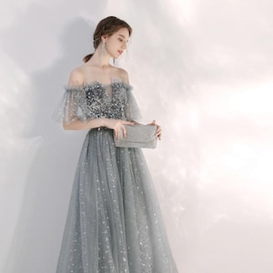 May include: A floor-length off-the-shoulder gown in a shimmering grey hue, featuring a bodice adorned with dense silver sequins and a tulle skirt embellished with a delicate star pattern, paired with a matching rectangular grey clutch bag.