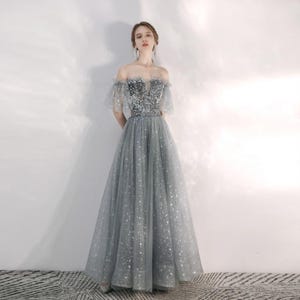 May include: A floor-length grey gown featuring an off-the-shoulder neckline and short tulle sleeves. The bodice is covered in silver sequins, while the full skirt is made of layered grey tulle fabric adorned with a subtle silver star pattern throughout.