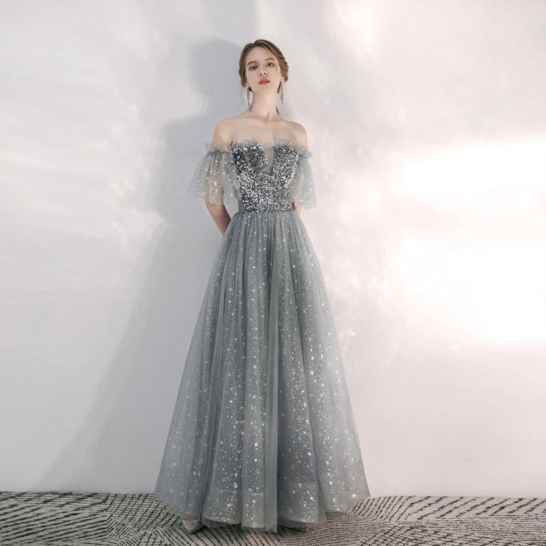 May include: A floor-length grey gown featuring an off-the-shoulder neckline and short tulle sleeves. The bodice is covered in silver sequins, while the full skirt is made of layered grey tulle fabric adorned with a subtle silver star pattern throughout.