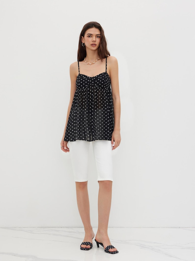 K&ouml;nnte beinhalten: Eine Frau tr&auml;gt ein schwarzes Polka-Dot-Camisole-Top mit d&uuml;nnen Tr&auml;gern und wei&szlig;e Shorts. Das Camisole hat einen Sweetheart-Ausschnitt. Sie tr&auml;gt au&szlig;erdem eine Perlenkette und schwarze Polka-Dot-Abs&auml;tze.