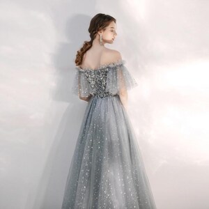 May include: A floor-length grey gown featuring an off-the-shoulder neckline with ruffled tulle sleeves. The bodice is embellished with silver sequins, while the skirt is crafted from layers of grey tulle adorned with a subtle star pattern.
