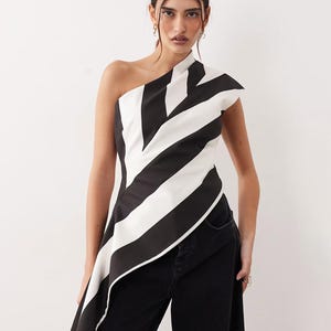 May include: A sleeveless one-shoulder top featuring bold, diagonal black and white stripes. The garment has a structured, asymmetrical hemline that extends longer on one side, creating a draped effect across the torso and waist area.
